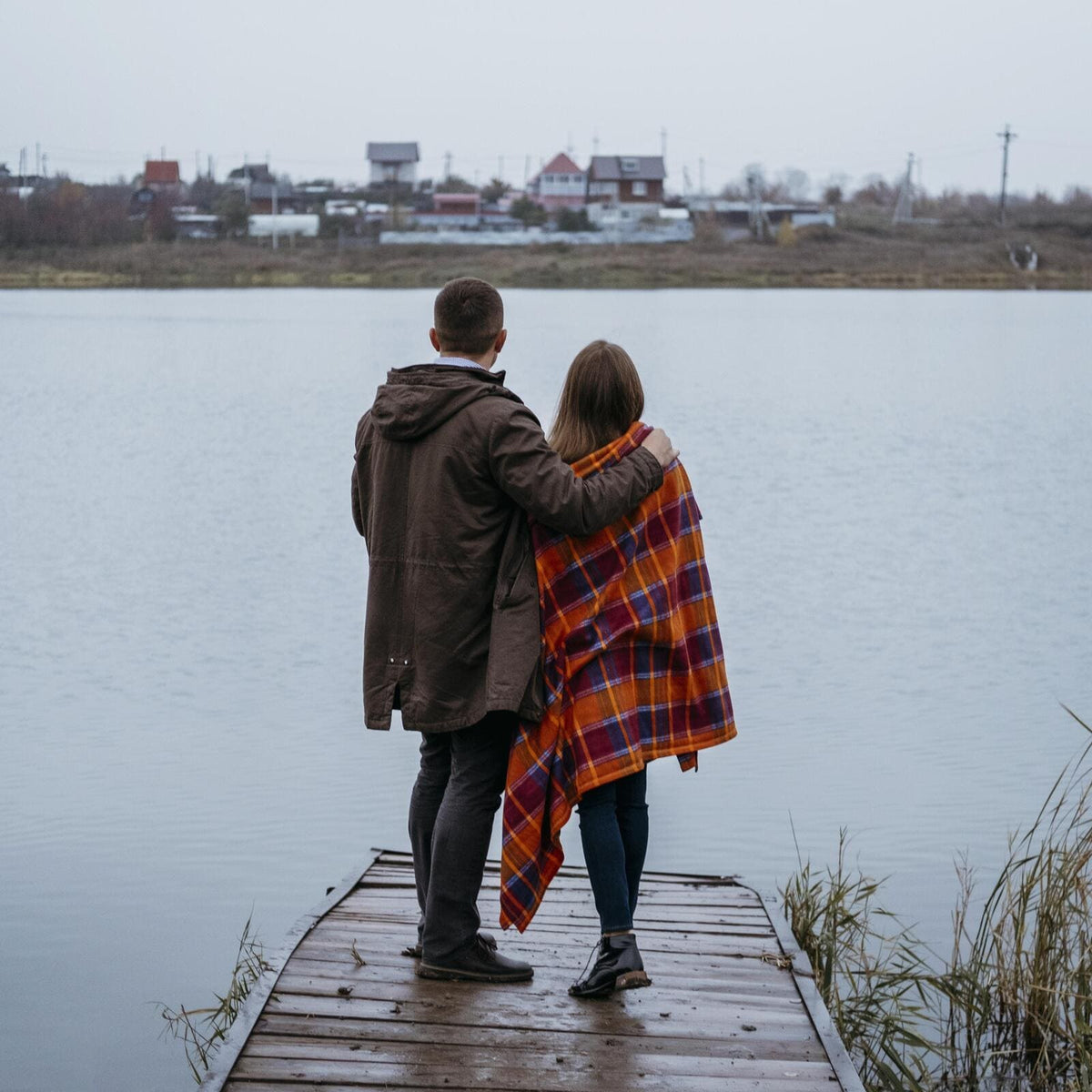Couple standing on a wooden jetty by a loch, wrapped in a tartan blanket on a grey autumn day